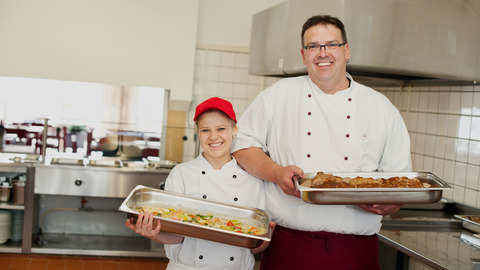 Eine Auszubildende und ein Ausbilder in der Küche. Sie halten jeweils einen Gastronormbehälter mit fertigem Essen in den Händen.