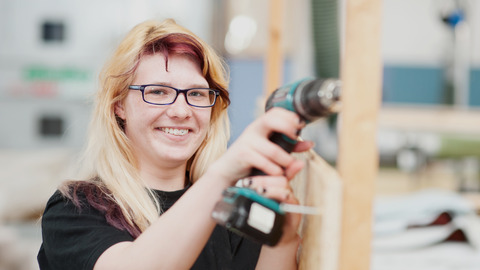 Eine Auszubildende bohrt mit einer Bohrmaschine in eine Holzkonstruktion.