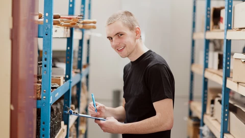 Ein Jugendlicher steht mit einem Klemmbrett zwischen Lagerregalen.