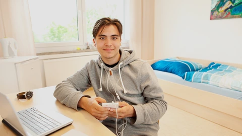 Ein Jugendlicher sitzt in einem Zimmer an einem Schreibtisch. Er hält ein Smartphone in der Hand. Vor ihm steht ein Laptop. Im Hintergrund ist ein Bett zu sehen.