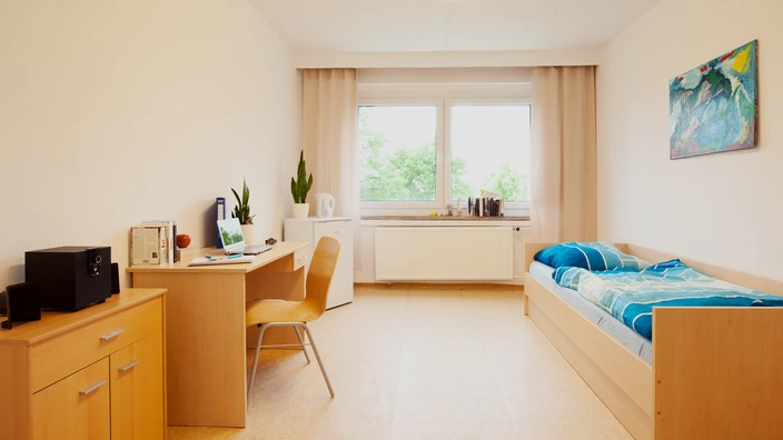 Einblick in ein Einzelzimmer im CJD Jugendgästehaus/Internat Ilmenau. Zu sehen sind eine Kommode, ein Schreibtisch, ein Kühlschrank und ein Bett.