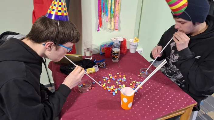 2 Jugendliche mit bunten Partyhütchen machen ein Partyspiel. Sie sitzen an einem Tisch und saugen bunte Schokolinsen mit einem langen Strohalm an, um sie in einem Glasgefäß zu sammeln.