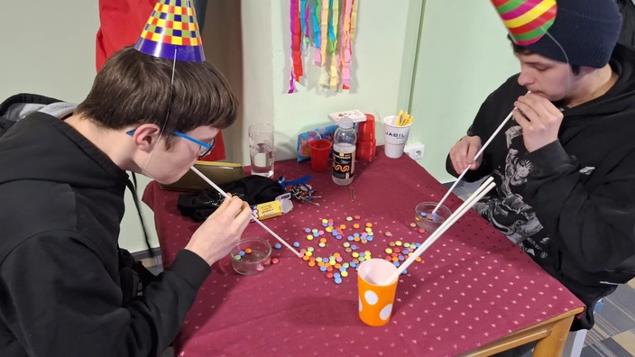 2 Jugendliche mit bunten Partyhütchen machen ein Partyspiel. Sie sitzen an einem Tisch und saugen bunte Schokolinsen mit einem langen Strohalm an, um sie in einem Glasgefäß zu sammeln.