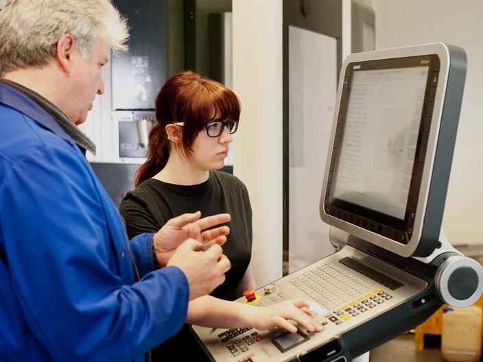 Ein Ausbilder und eine Auszubildende stehen vor einem Eingabegerät mit großem Monitor.