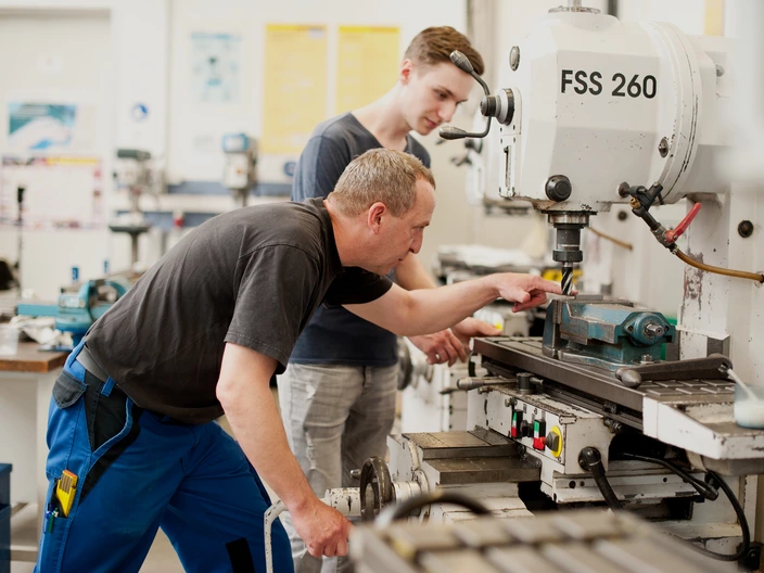 Ein Ausbilder erklärt einem Auszubildenden etwas an einer Maschine im Metallbereich.